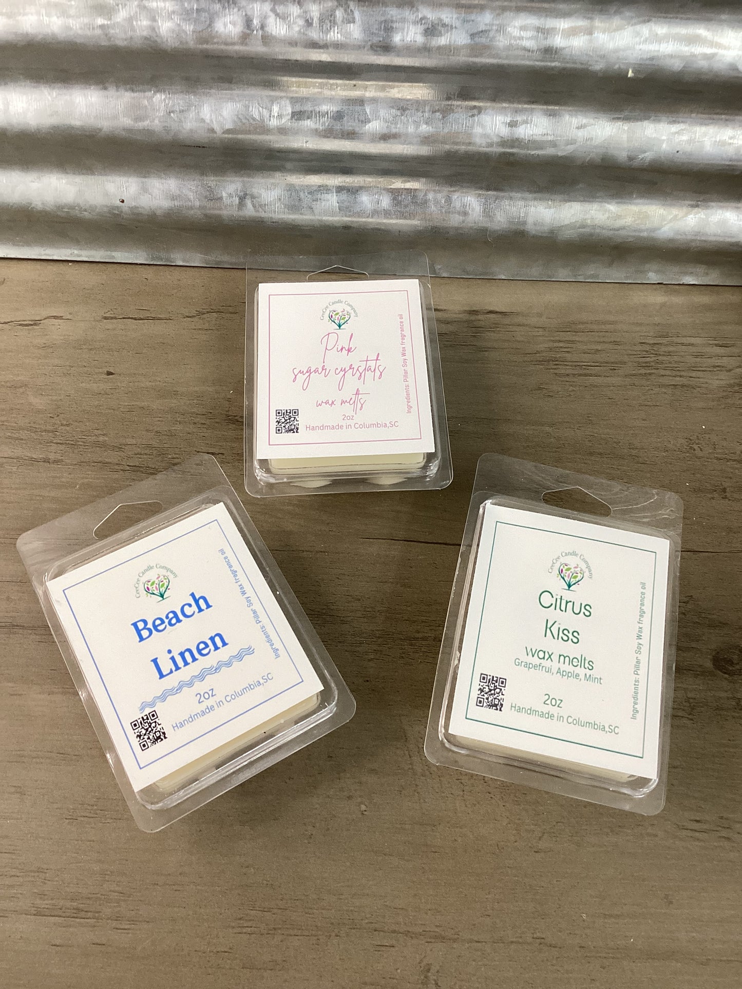 The Build Your Own Wax Melts Bundle from CeeCee Candle Company features Beach Linen, Pink Sugar Crystals, and Citrus Kiss packs displayed on wood with a metal backdrop—customize your perfect scent set for any space.