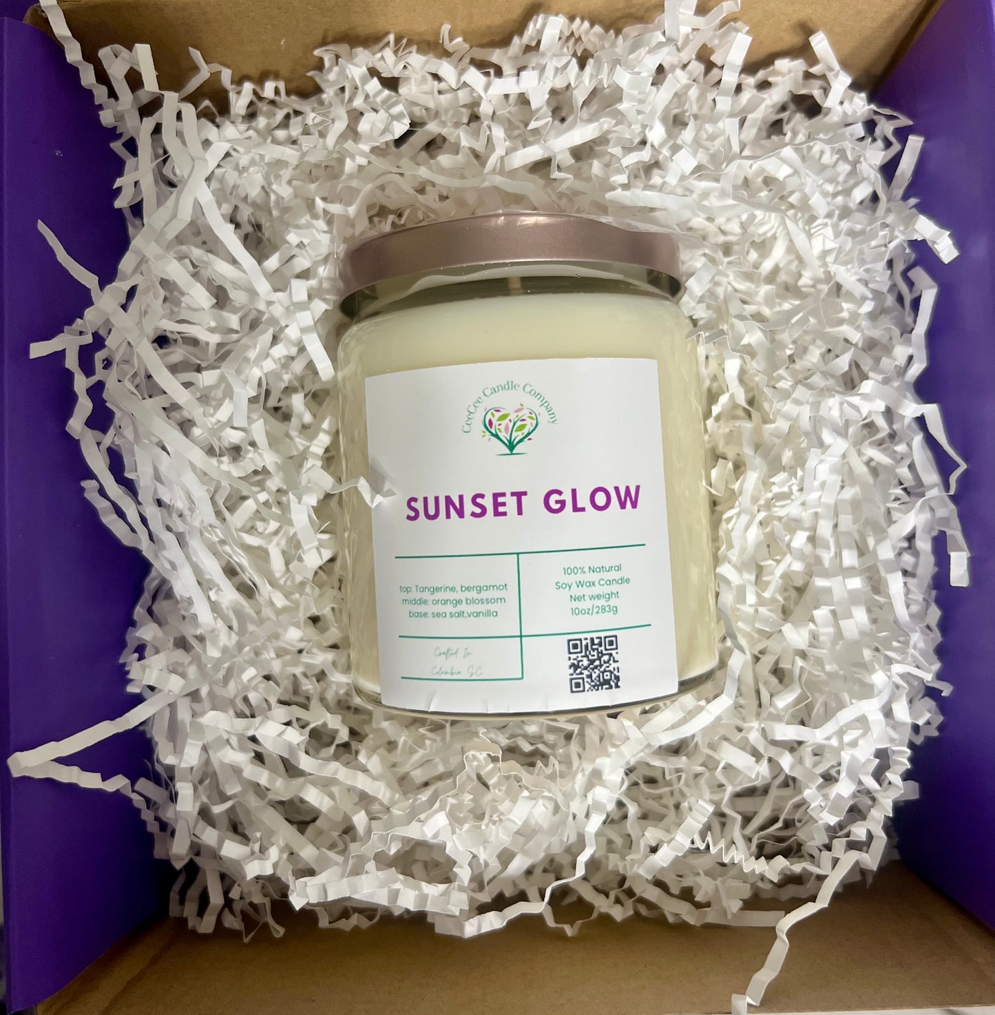 A CeeCee Candle Company Signature Apothecary Collection soy candle sits in a box of white crinkle paper. It features a silver lid, pastel label with QR code, premium fragrance oil notes, and highlights its natural soy wax.