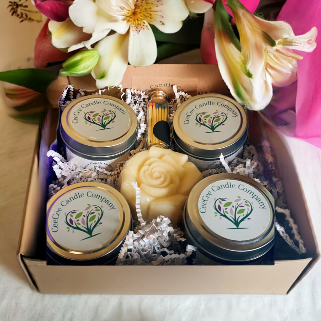 The CeeCee Candle Company Gift Box features four tin soy candles, a rose-shaped candle, matches, and decorative shredded paper, all in eco-friendly packaging by CeeCee Candle Company. Flowers are shown in the background.