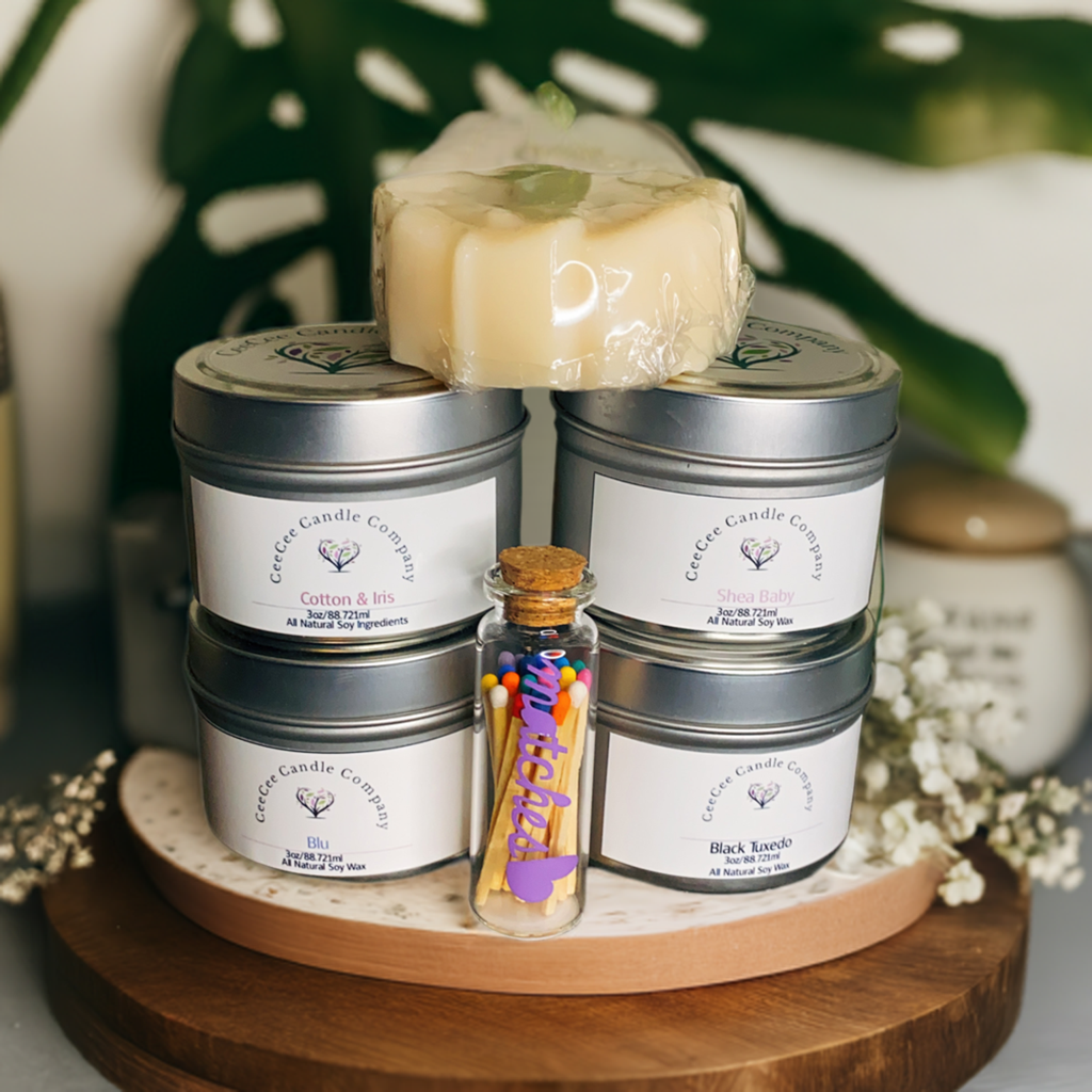 The CeeCee Candle Company Gift Box by CeeCee Candle Company features four soy candles in tins, a handmade soap bar, and a glass vial of matches on a wooden tray—perfect for self-care, with green leaves and white flowers in the background.
