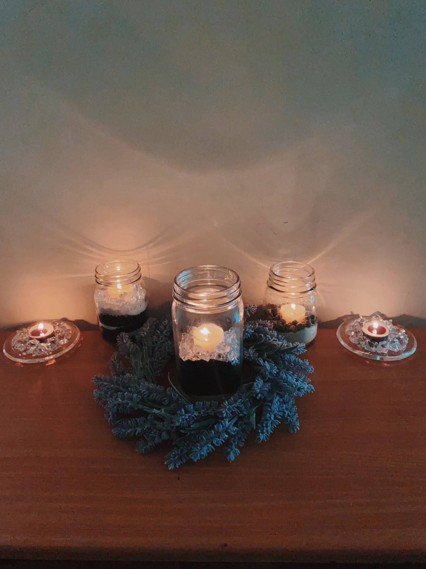 Three mason jars with candles sit on a wooden surface, the center one encircled by a blue pine wreath. On either side, CeeCee Candle Company Soy Wax Tea Light Candles in glass holders add an eco-friendly, cozy glow.