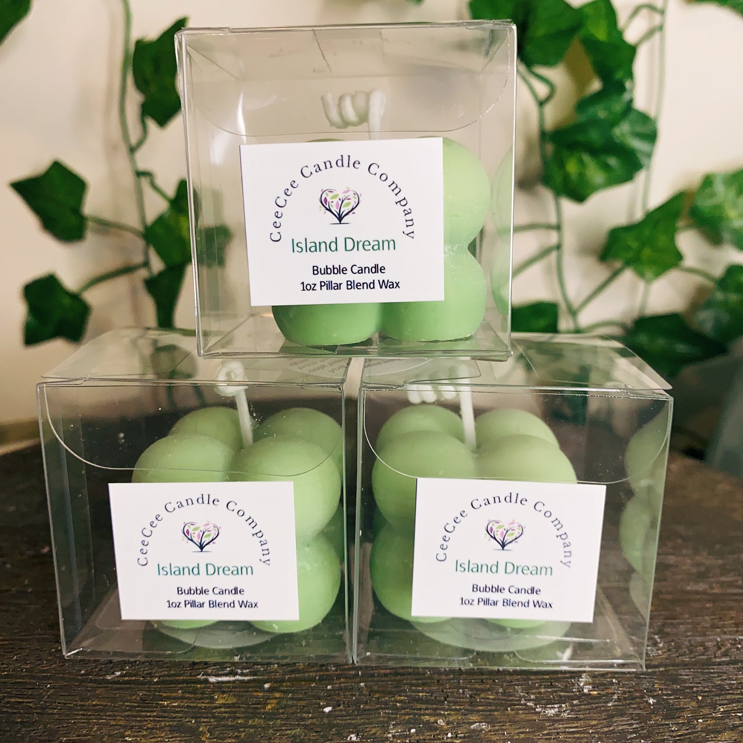 Three light green Bubble Candles with glass dishes from CeeCee Candle Company are stacked in a pyramid. Each candle adds lush fragrance, echoing the blurred green leaves in the background.
