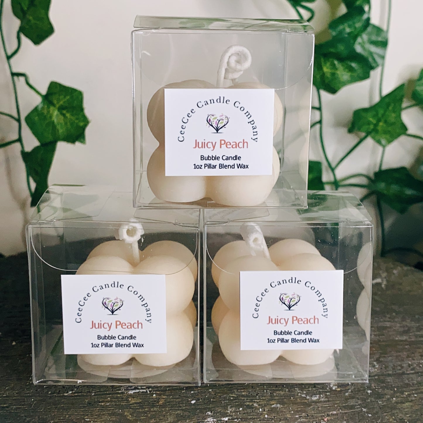 Three cream-colored Bubble Candles with glass dishes from CeeCee Candle Company are displayed in clear plastic boxes, stacked pyramid-style against green ivy, ready to fill your space with delightful fragrance oils.