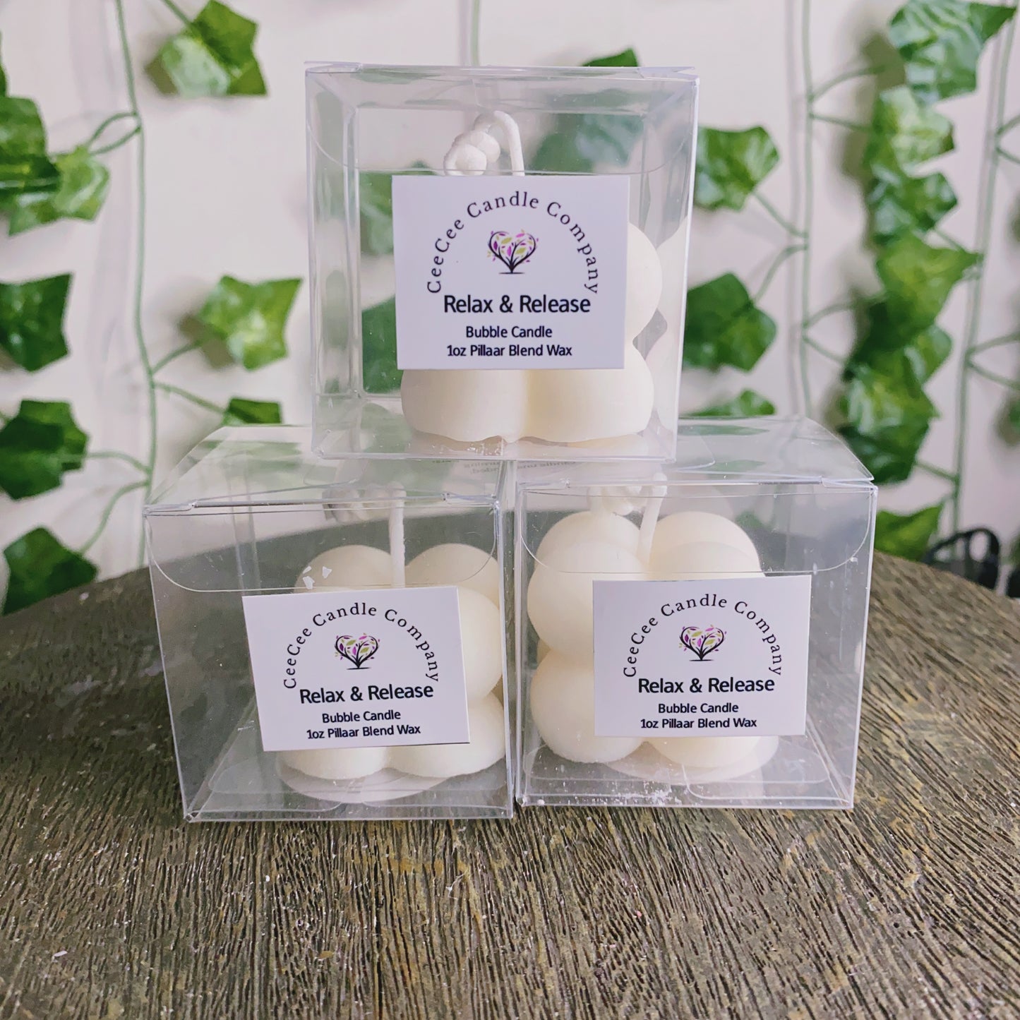 Three white Bubble Candles with glass dishes from CeeCee Candle Company are stacked on a rustic wooden surface amid green plants—perfect for pairing with your favorite fragrance oils.
