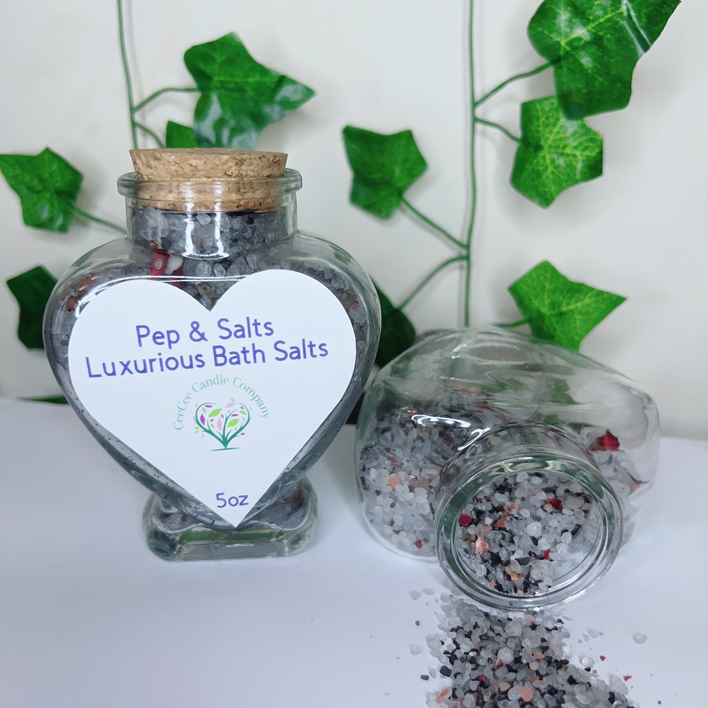 Two glass jars of Pep and Salts Luxurious Bath Salts by CeeCee Candle Company are shown—one upright and heart-shaped, labeled with Himalayan pink salt, while the other lies sideways spilling salts. Green ivy leaves decorate the background.