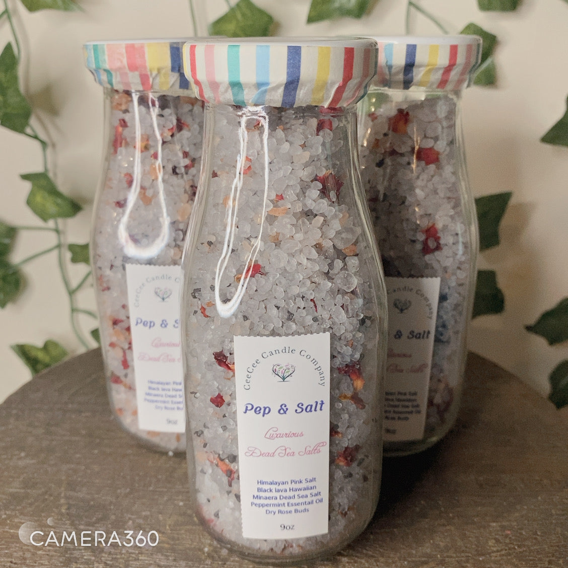 Three Pep and Salts Luxurious Bath Salts by CeeCee Candle Company, in glass jars with colorful striped lids and ingredient labels, are displayed in front of leafy green decor.