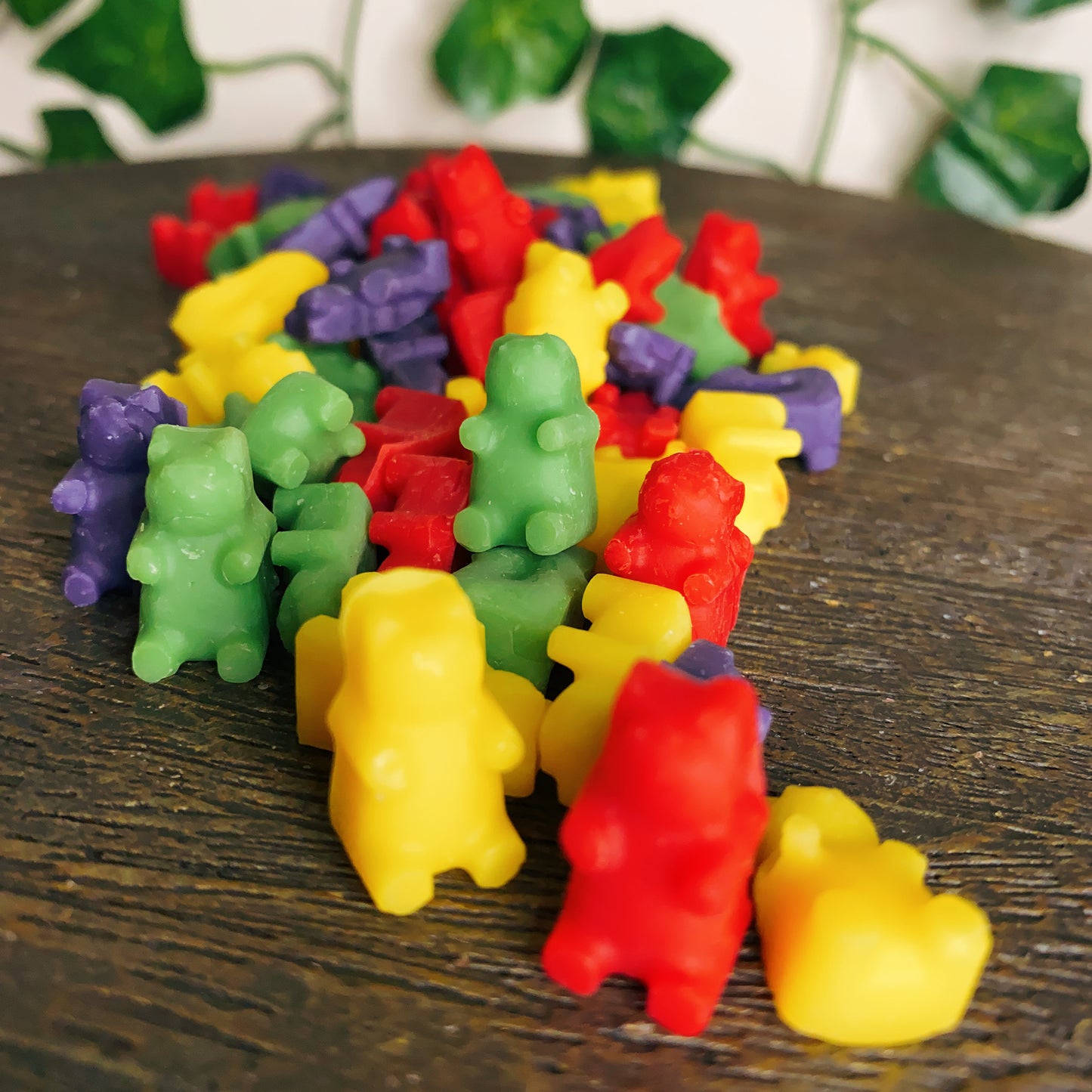 A pile of colorful Moroccan Cashmere Gummy Bear Wax Melts by CeeCee Candle Company sits on a wooden surface, with blurred green leaves in the background, evoking a relaxing and vibrant atmosphere.