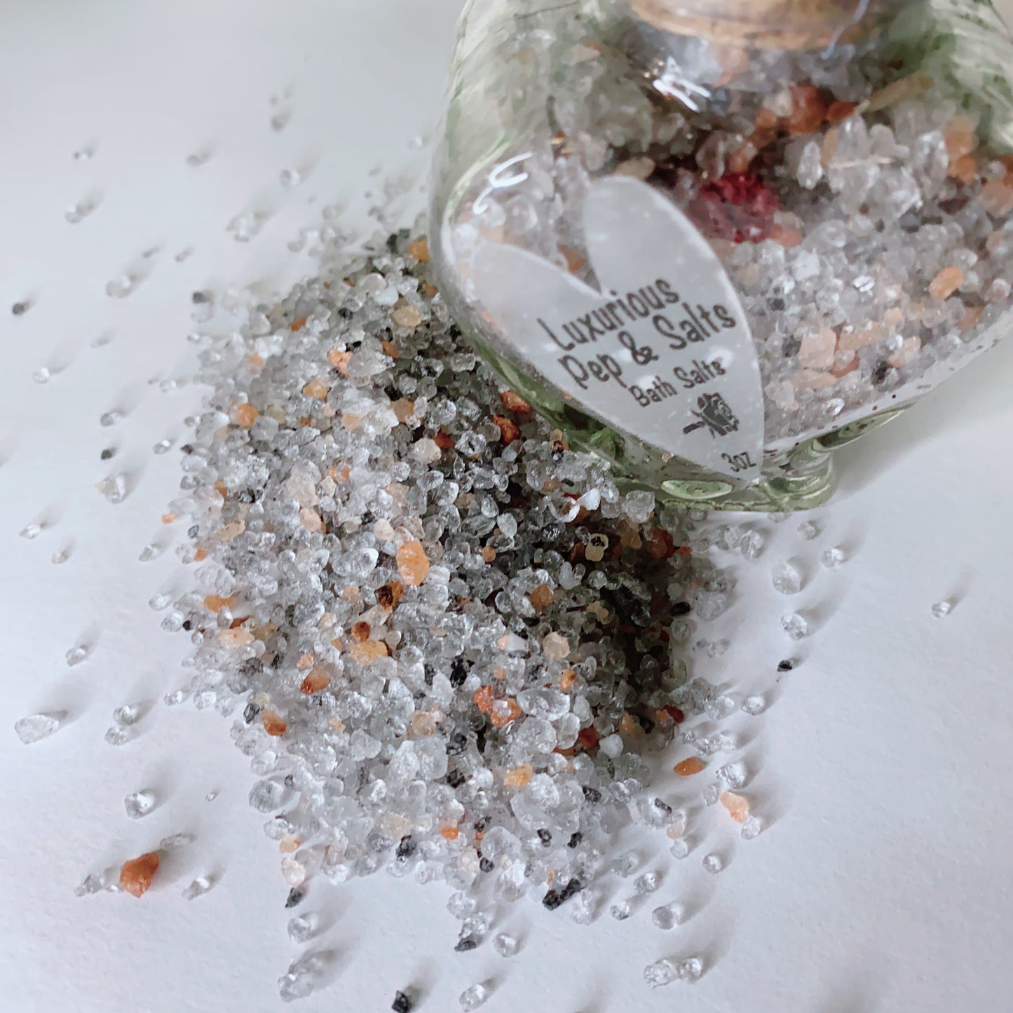 A glass jar of Pep and Salts Luxurious Bath Salts by CeeCee Candle Company spills vibrant crystals onto a white surface. Made with Himalayan pink salt, these bath salts offer a luxurious aromatherapy experience.