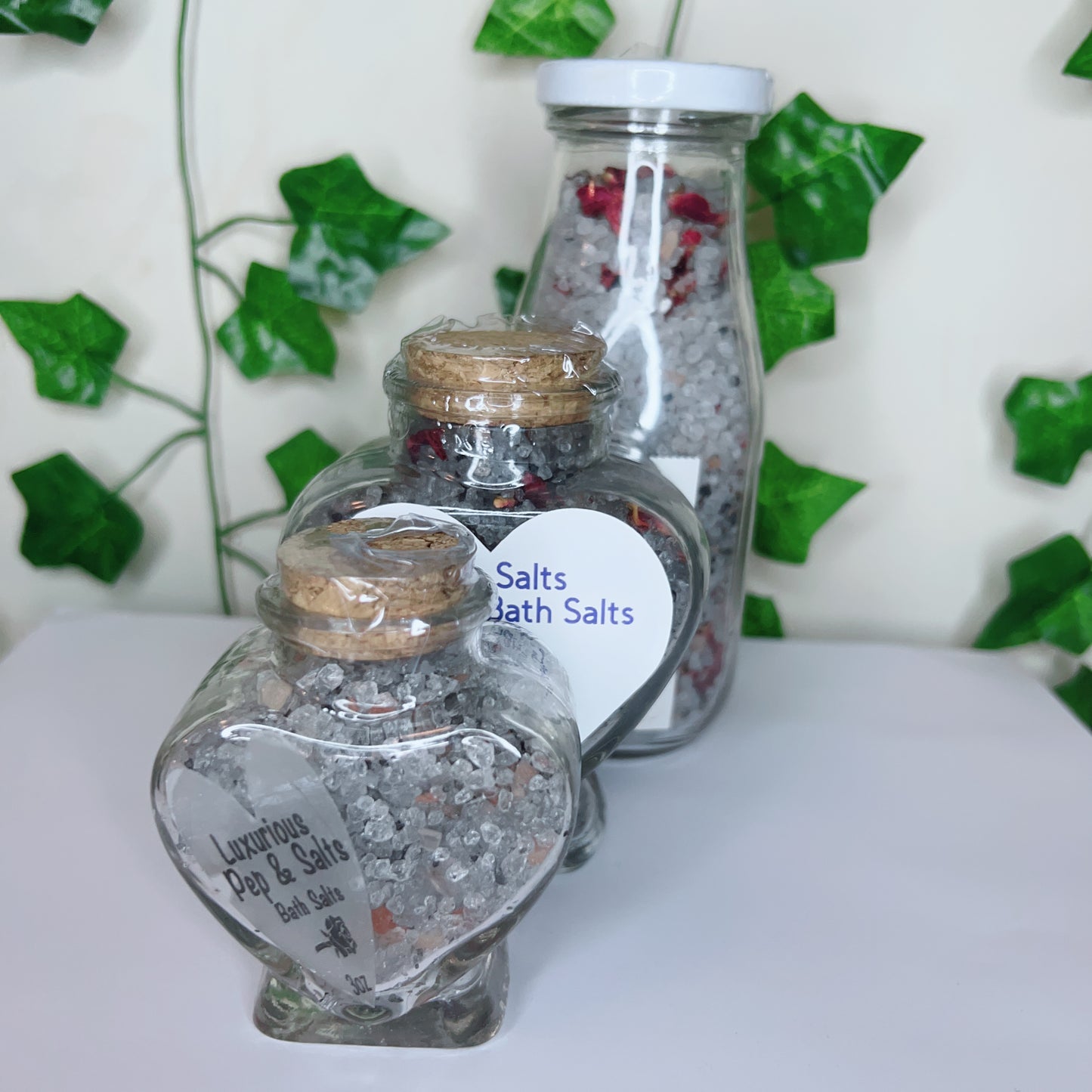 Three jars of Pep and Salts Luxurious Bath Salts by CeeCee Candle Company, featuring Himalayan pink salt, are displayed on a white surface. Heart-shaped and tall bottles with cork lids sit beneath green faux leaves on the wall.