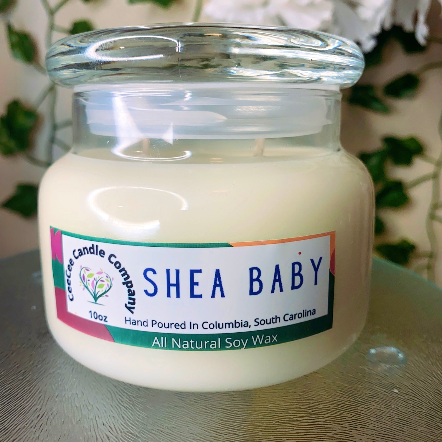 A glass jar candle labeled "Shea Baby" by Cece Candle Company sits on a table. The label notes it is hand-poured in Columbia, South Carolina and made from all natural soy wax.