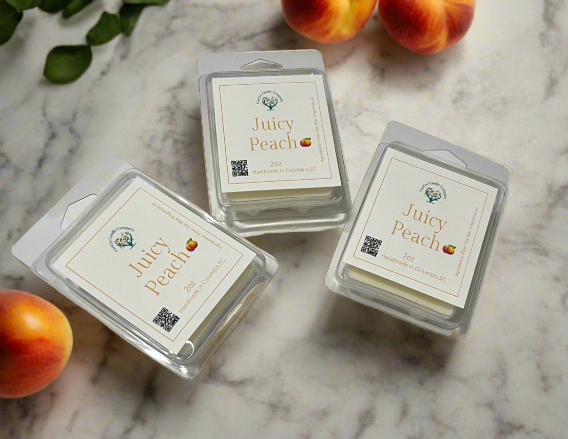 Three packs of "Juicy Peach" wax melts on a marble surface, with two peaches and green leaves nearby. The label reads "Handmade in Columbia, SC" with a peach emoji.