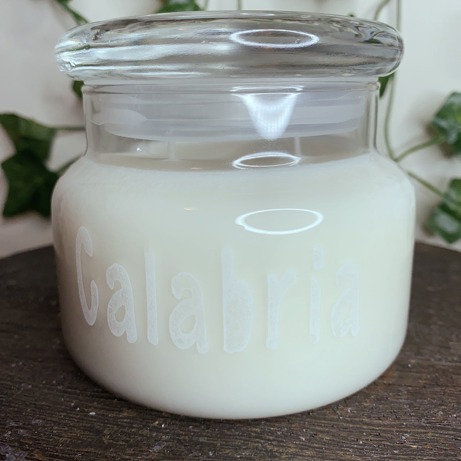 A glass jar candle with white wax and the word "Calabria" etched on the front, sitting on a wooden surface with green leaves in the background.