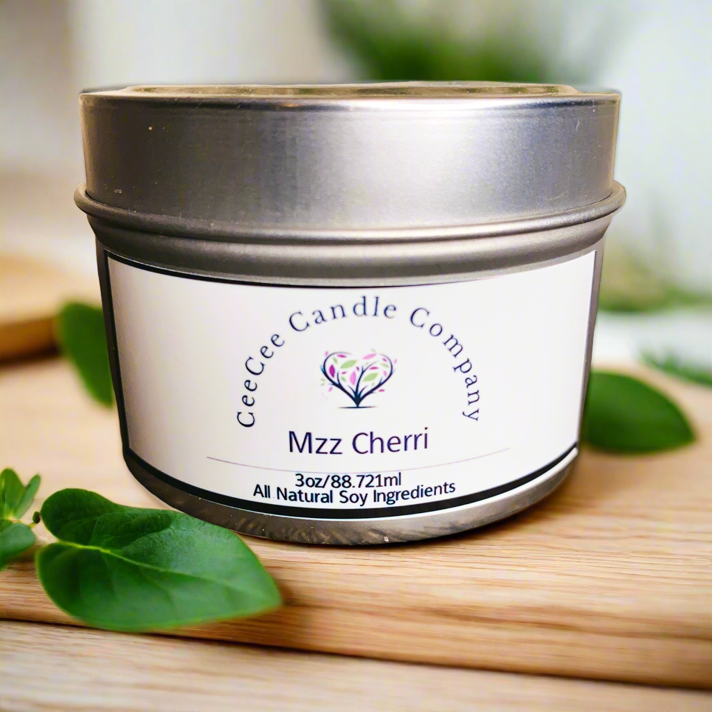 The CeeCee Candle Company Travel Size Soy Candle, 3oz tin, features the "Mzz Cherri" scent made from all-natural soy wax and delivers a mood-boosting fragrance. Displayed on a wooden surface with fresh green leaves.