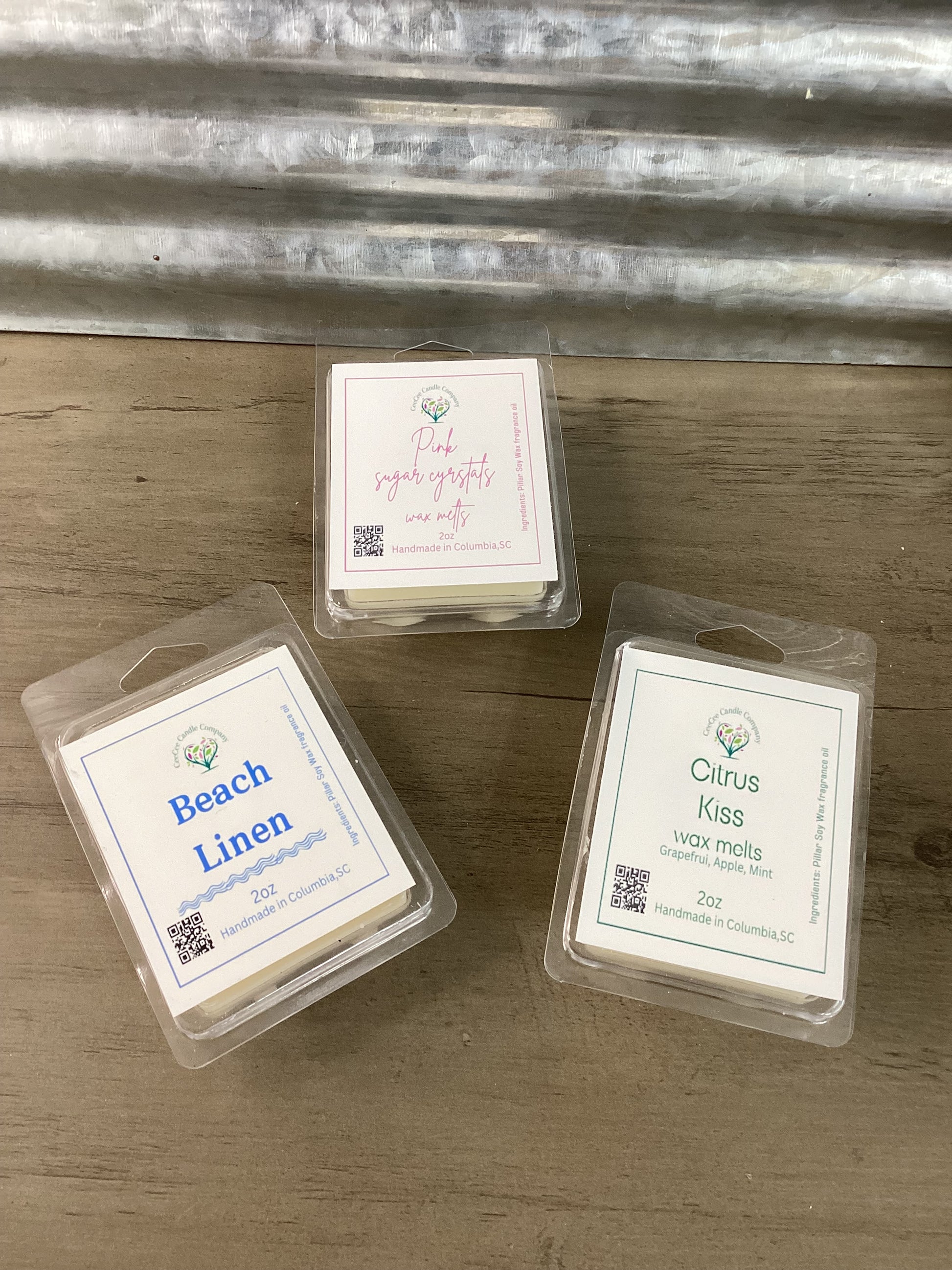 The Build Your Own Wax Melts Bundle from CeeCee Candle Company features Beach Linen, Pink Sugar Crystals, and Citrus Kiss packs displayed on wood with a metal backdrop—customize your perfect scent set for any space.