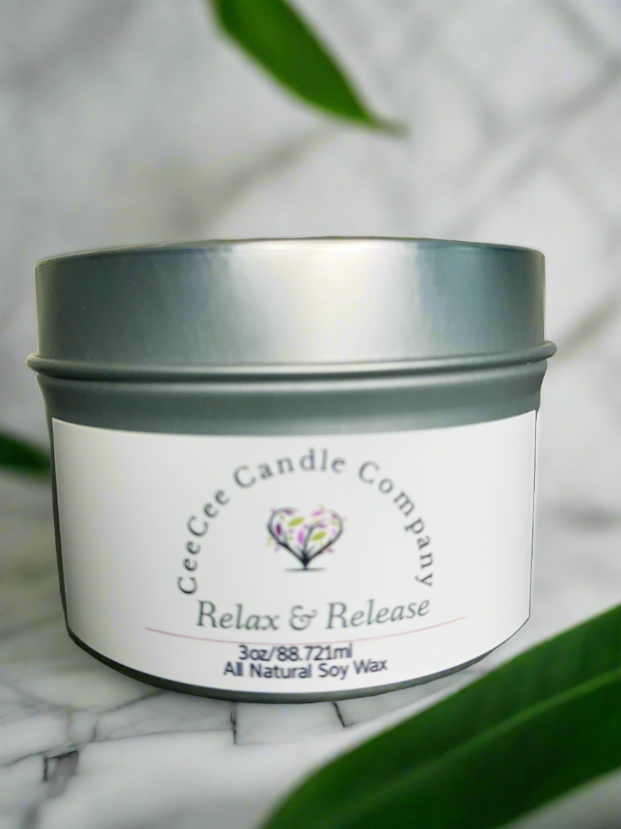 A silver tin Travel Size Soy Candle by CeeCee Candle Company rests on a white surface, with blurred green leaves in the foreground.