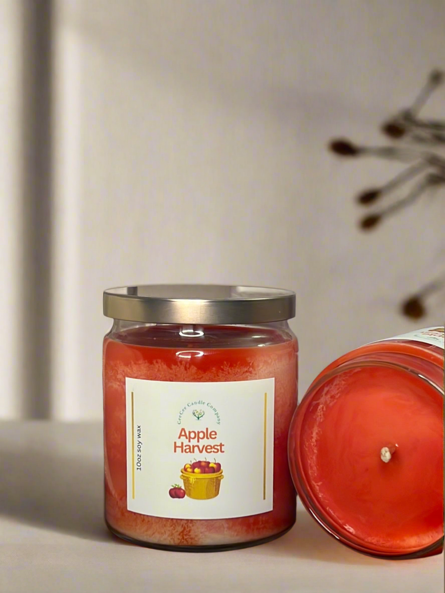 A CeeCee Candle Company Signature Apothecary Collection jar candle labeled "Apple Harvest," featuring apples and a basket, sits on a light surface with another similar soy wax candle nearby. The softly lit background is slightly out of focus.