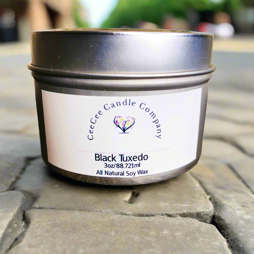 A small CeeCee Candle Company Travel Size Soy Candle sits on cobblestone pavement, with blurred greenery and buildings in the background.