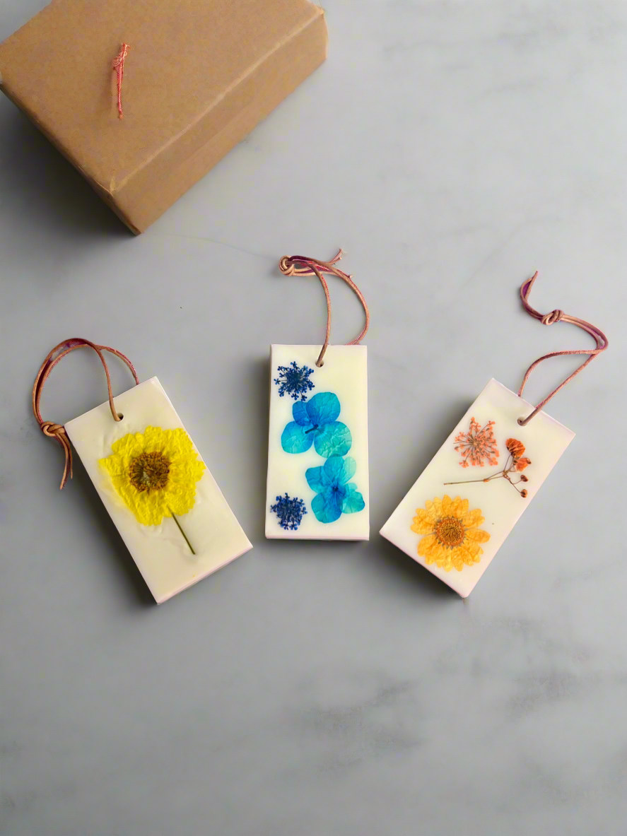 Three rectangular Wax Sachets by CeeCee Candle Company, made from soy wax and decorated with dried flowers and twine loops, are displayed on a light surface beside a closed brown box. Each sachet has a distinct, colorful floral design.