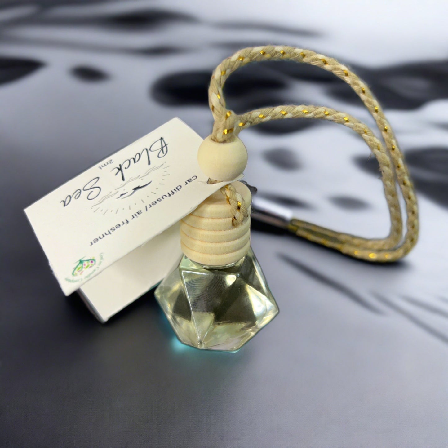 A CeeCee Candle Company Car diffuser with a wooden cap and rope, made from natural materials, rests beside its white box on a gray surface. The glass bottle holds a light liquid that releases a refreshing scent.