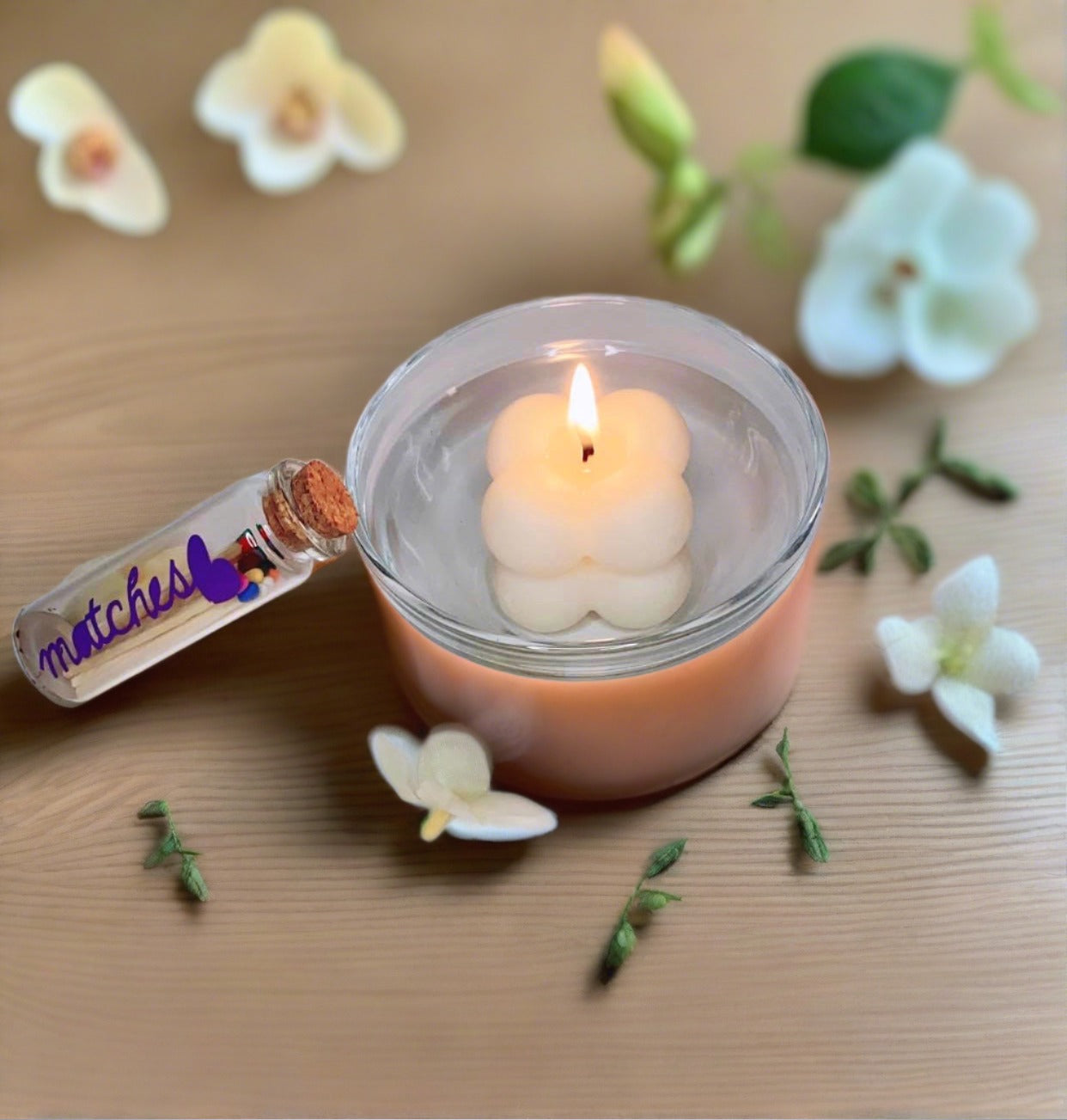 A lit Bubble Candle with glass dish by CeeCee Candle Company floats in water, surrounded by white flowers and green leaves. A small vial labeled "matches" sits nearby, creating an inviting setting for gentle light and fragrance oils.