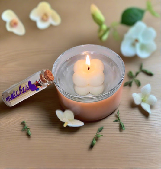 A lit Bubble Candle with glass dish by CeeCee Candle Company floats in water, surrounded by white flowers and green leaves. A small vial labeled "matches" sits nearby, creating an inviting setting for gentle light and fragrance oils.