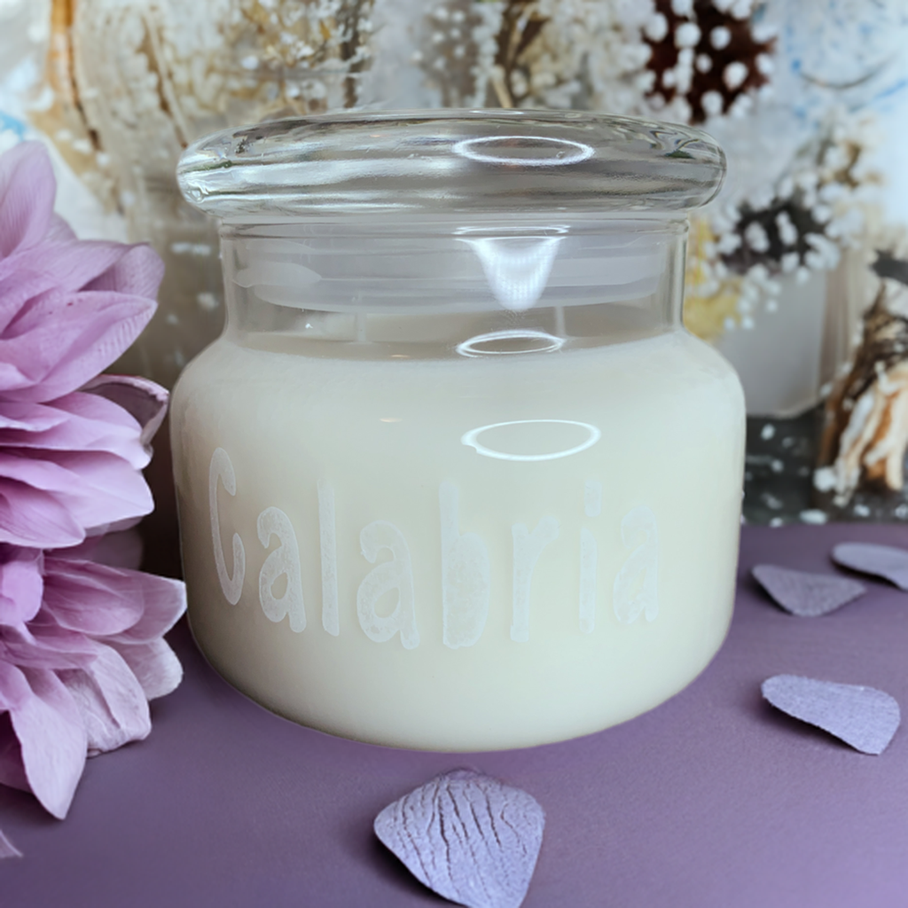 A glass jar soy candle from CeeCee Candle Company’s Custom Gifts collection sits on a purple surface with a pink flower and purple petals, set against a blurred botanical background—ideal for natural scent lovers.