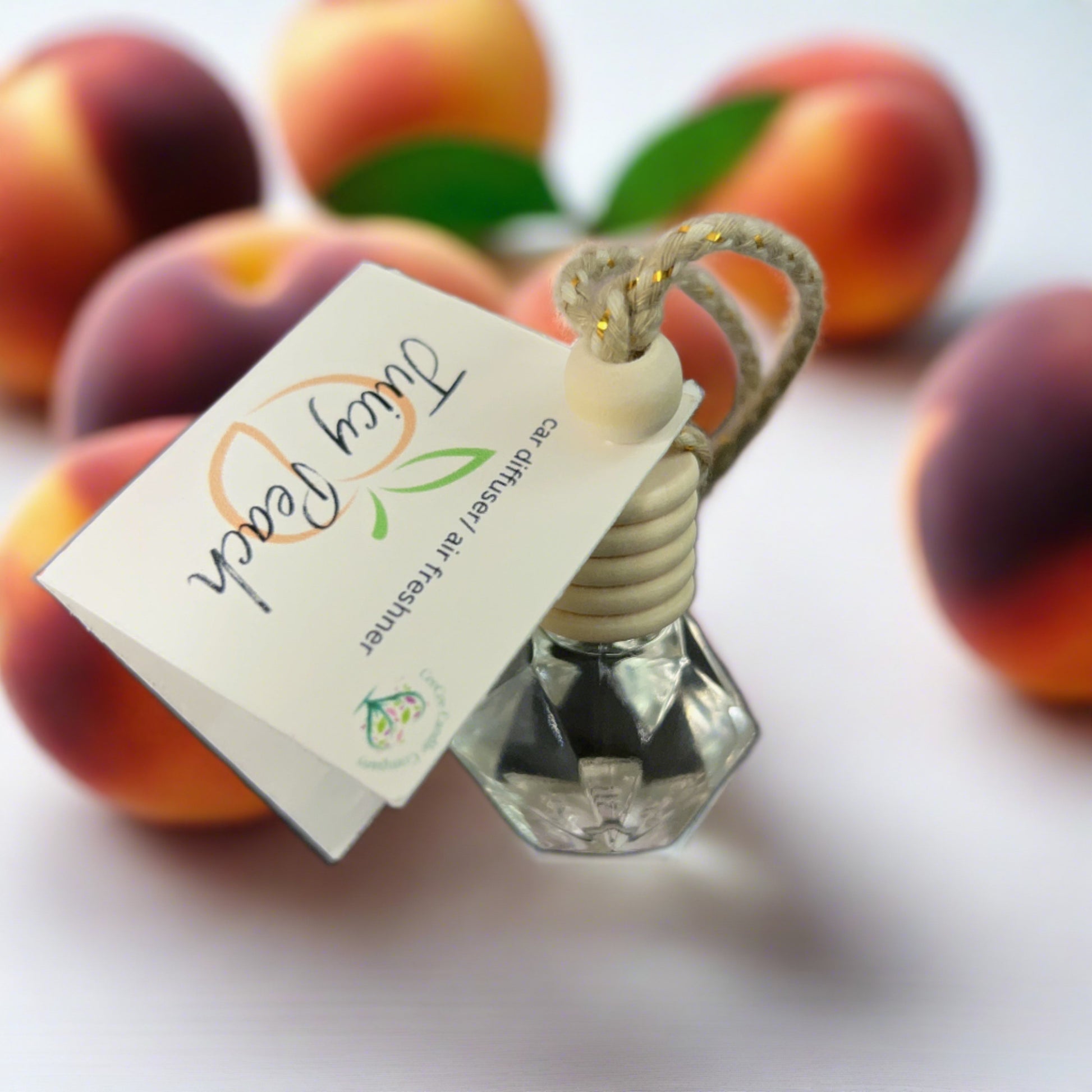 A small CeeCee Candle Company Car Diffuser with a rope labeled "Juicy Peach" rests on a white surface, surrounded by peaches, offering a refreshing natural aroma.
