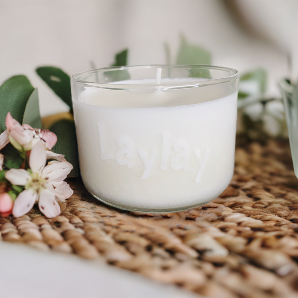 A white soy candle from CeeCee Candle Company’s Custom Gifts line, in a glass container embossed with "Laylay," sits on a woven mat among pink and white flowers and green leaves, filling the space with gentle, natural scents.