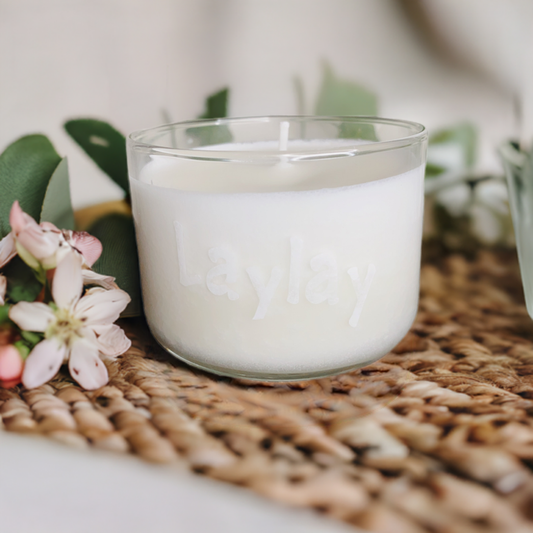 A white soy candle from CeeCee Candle Company’s Custom Gifts line, in a glass container embossed with "Laylay," sits on a woven mat among pink and white flowers and green leaves, filling the space with gentle, natural scents.