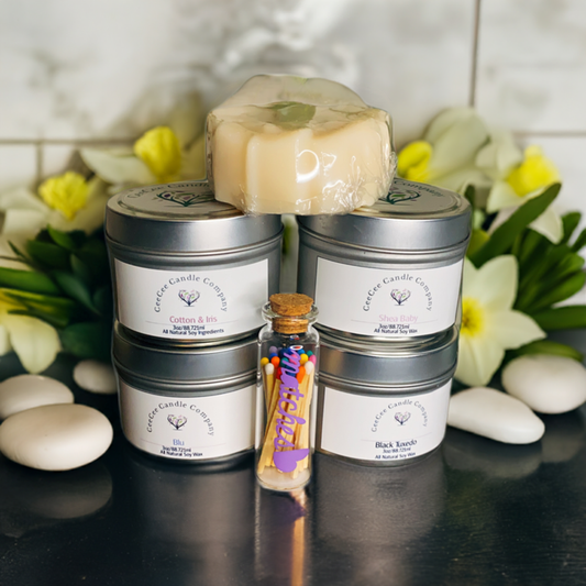A display of four tin soy candles stacked with a wrapped soap bar on top, a small corked glass bottle of colorful matches in front, and delicate white flowers and stones—perfect for the Floral Escape Candle Gift Box by CeeCee Candle Company.