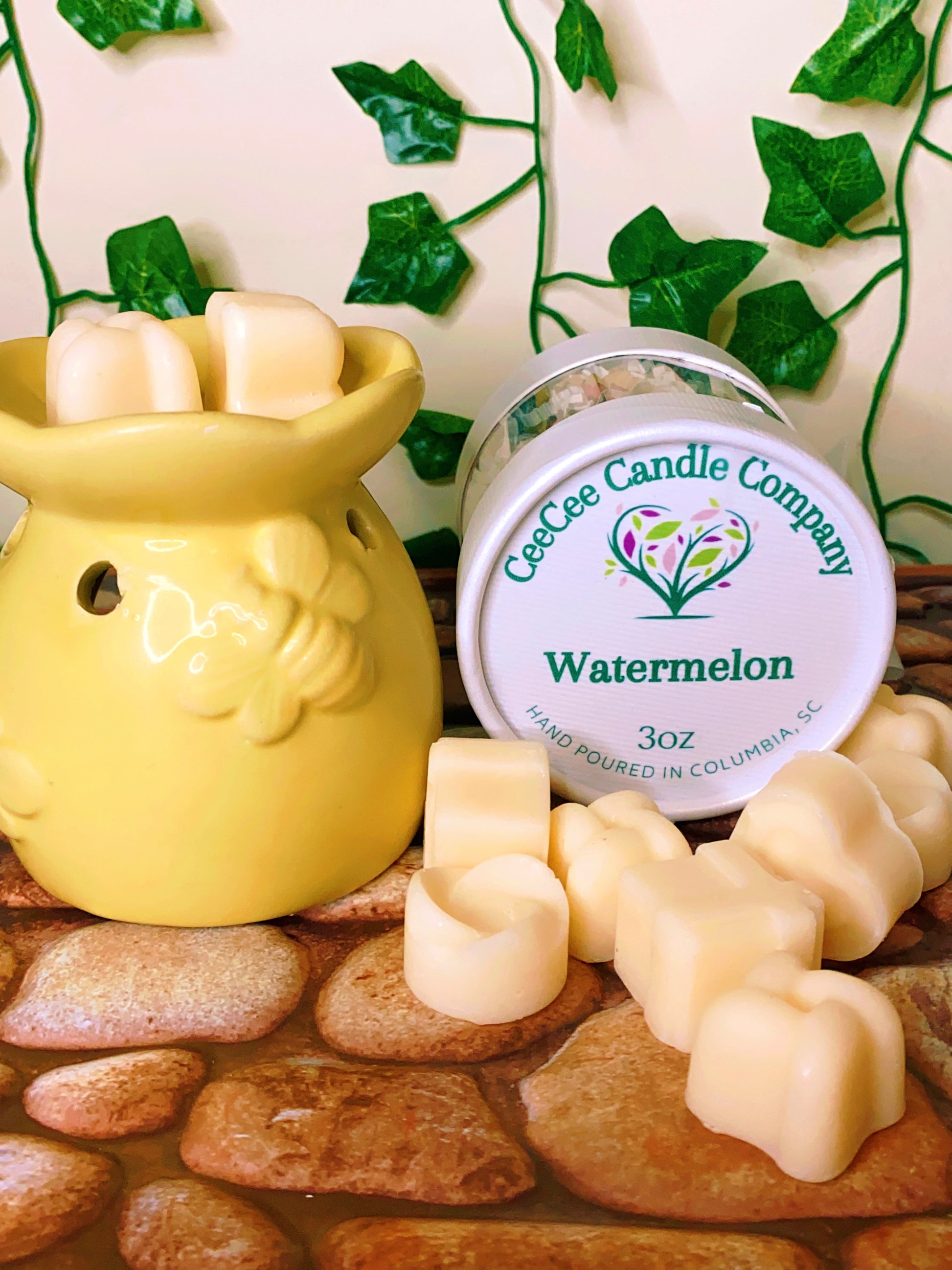 A yellow wax warmer holds creamy Wax Melts Gift Box by CeeCee Candle Company, with heart-shaped melts scattered on a stone-patterned surface and green vines in the background.
