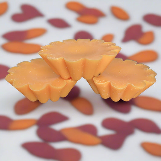 Three orange, flower-shaped Candle Tart Melts from CeeCee Candle Company are stacked in a small pyramid against a backdrop of orange and brown petal-like shapes. The bright setting highlights their smooth, slightly glossy texture.