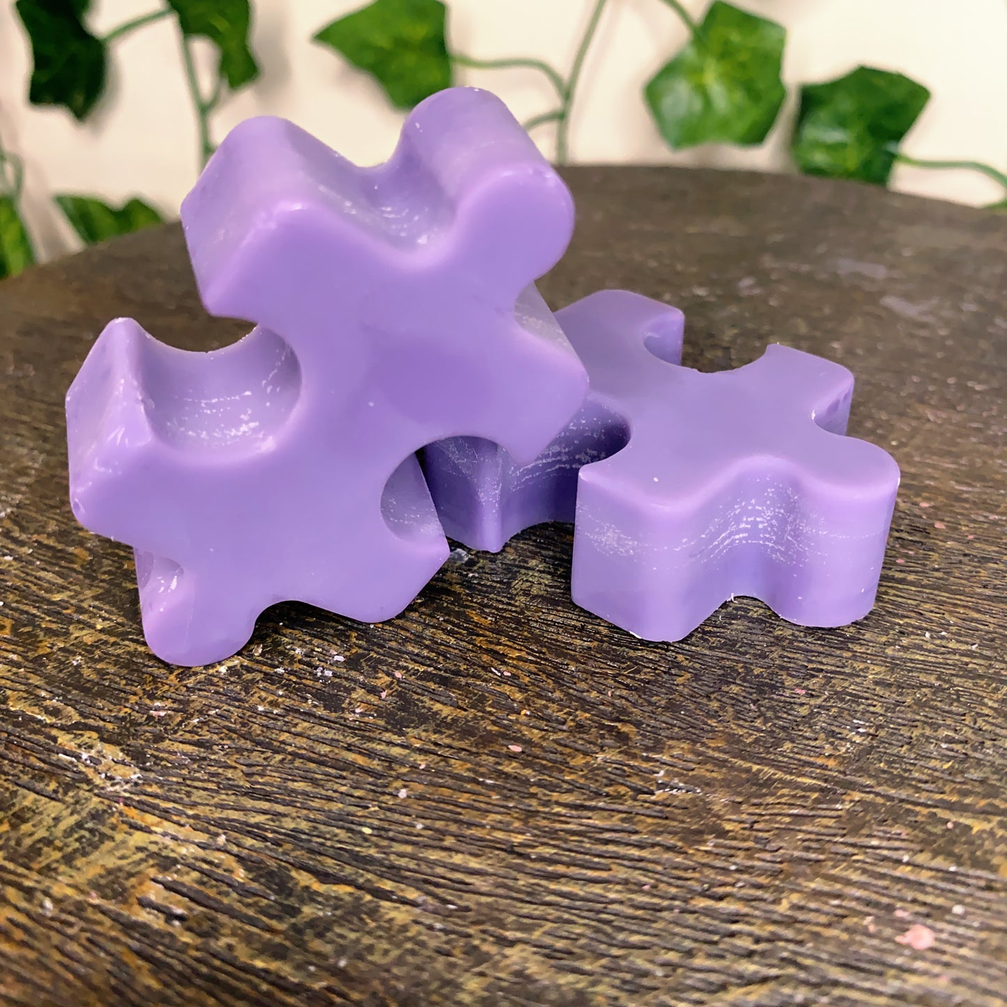 Two purple Puzzle Pieces Wax Melts from CeeCee Candle Company sit on a wooden surface, with blurred leafy green vines in the background.