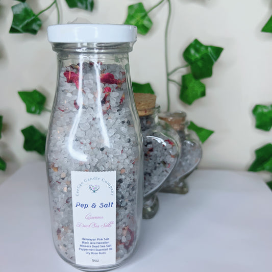 A clear glass bottle of Pep and Salts Luxurious Bath Salts by CeeCee Candle Company, filled with coarse Himalayan pink salt and dried rose petals, stands before ivy-wrapped walls and heart-shaped jars in the background.