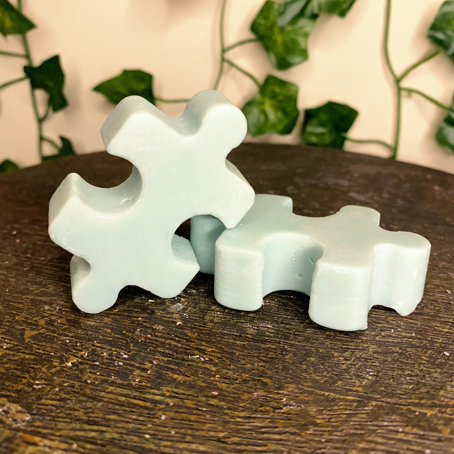 Two light blue Puzzle Pieces Wax Melts by CeeCee Candle Company sit on a dark wooden surface, with blurred green leafy vines in the background, adding charm and inviting fragrance to the scene.