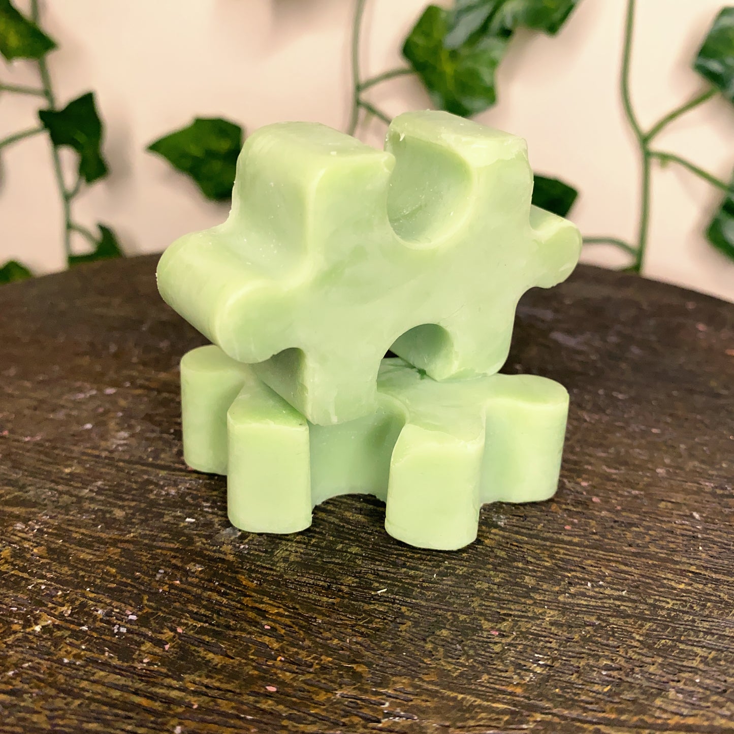 A green Puzzle Pieces Wax Melt by CeeCee Candle Company rests on a wooden surface, while leafy vines are softly blurred in the background.