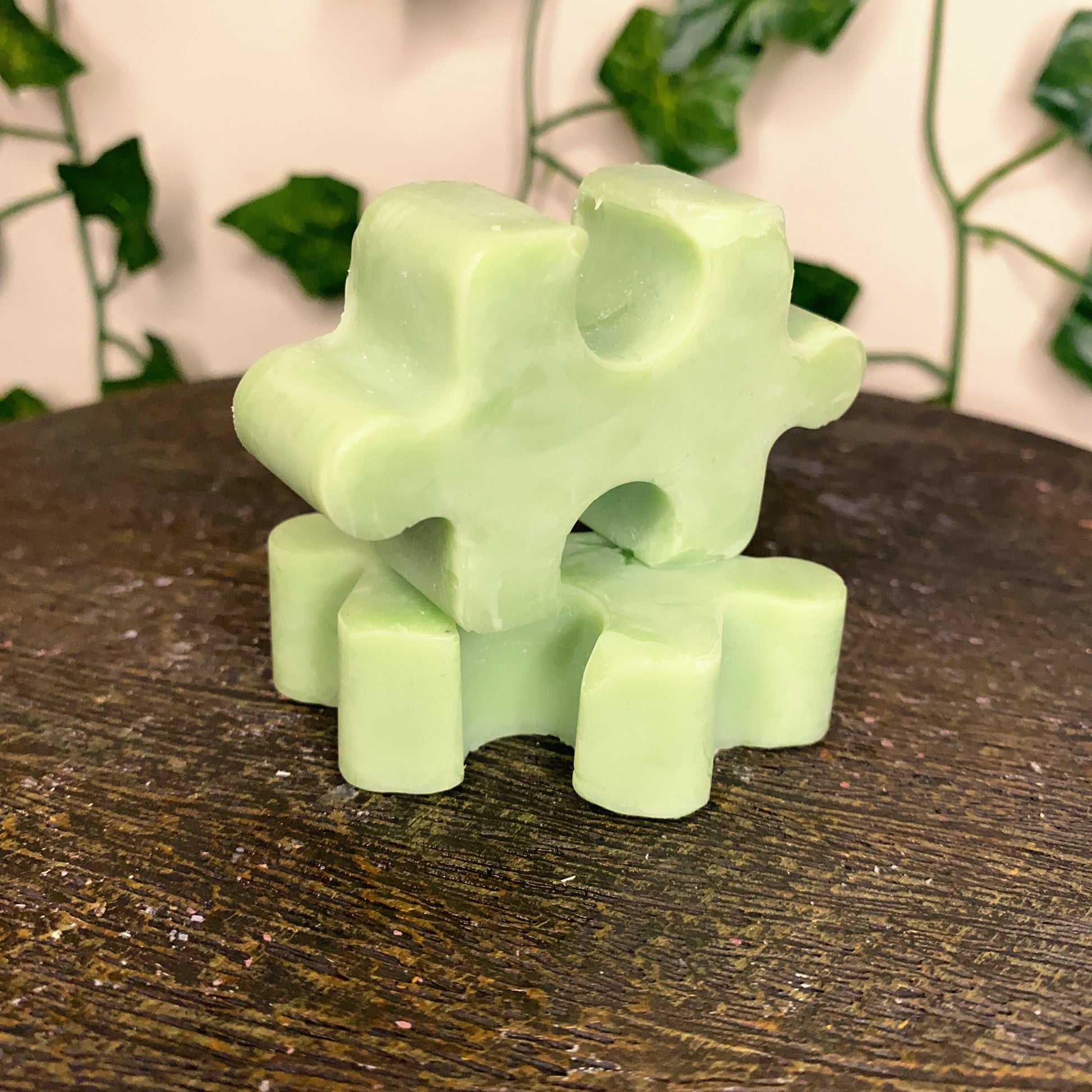 A green Puzzle Pieces Wax Melt by CeeCee Candle Company rests on a wooden surface, while leafy vines are softly blurred in the background.