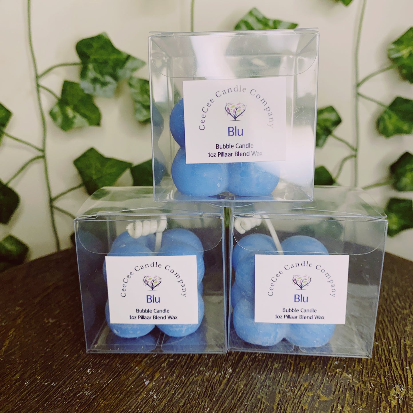 Three blue "Bubble Candle with glass dish" by CeeCee Candle Company are shown in clear boxes, stacked in a pyramid. Each box is labeled and green ivy leaves hang in the background.