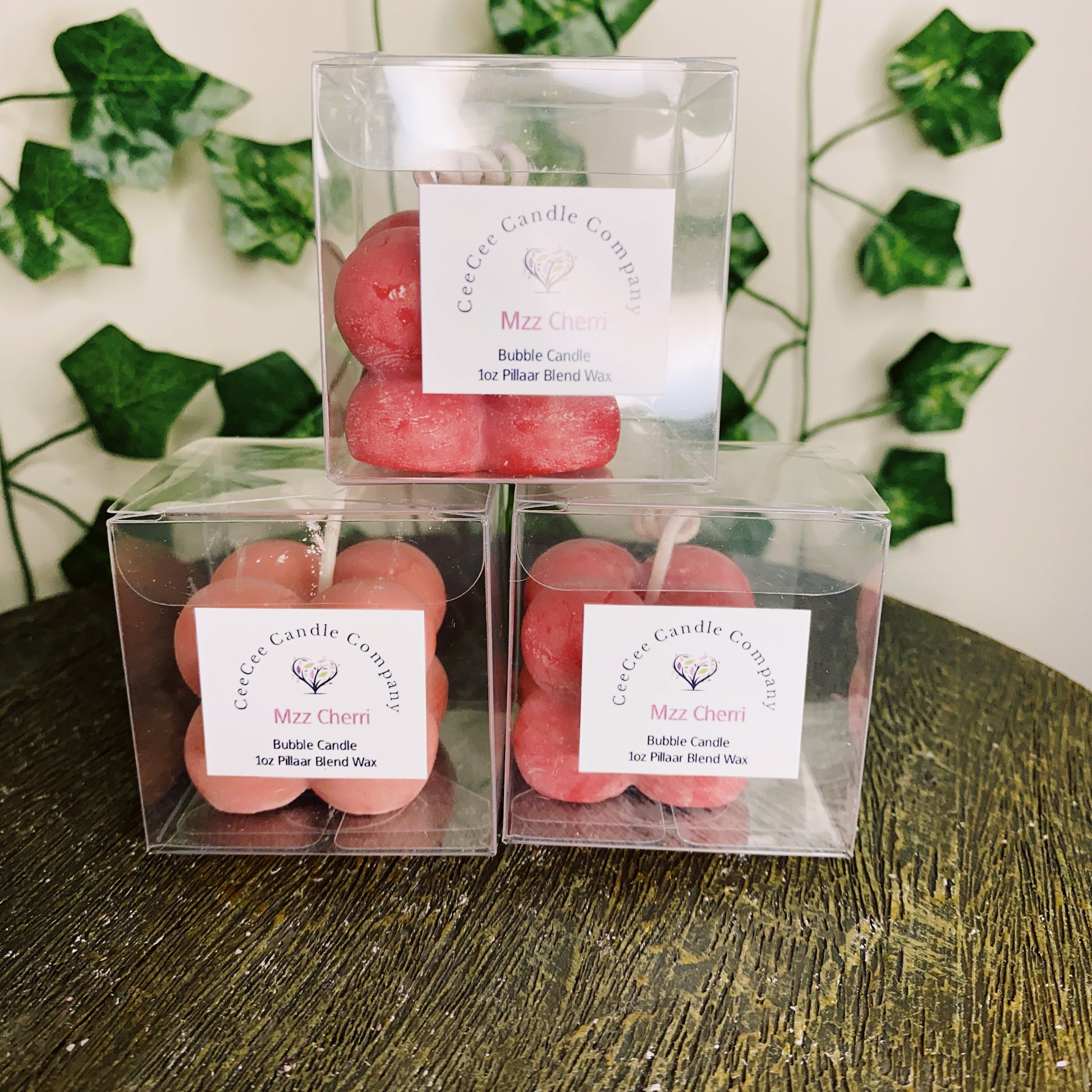 Three red, bubble-shaped Bubble Candles with glass dishes by CeeCee Candle Company are individually boxed with white labels and displayed on a wooden surface with greenery—ideal for use in a burning dish.