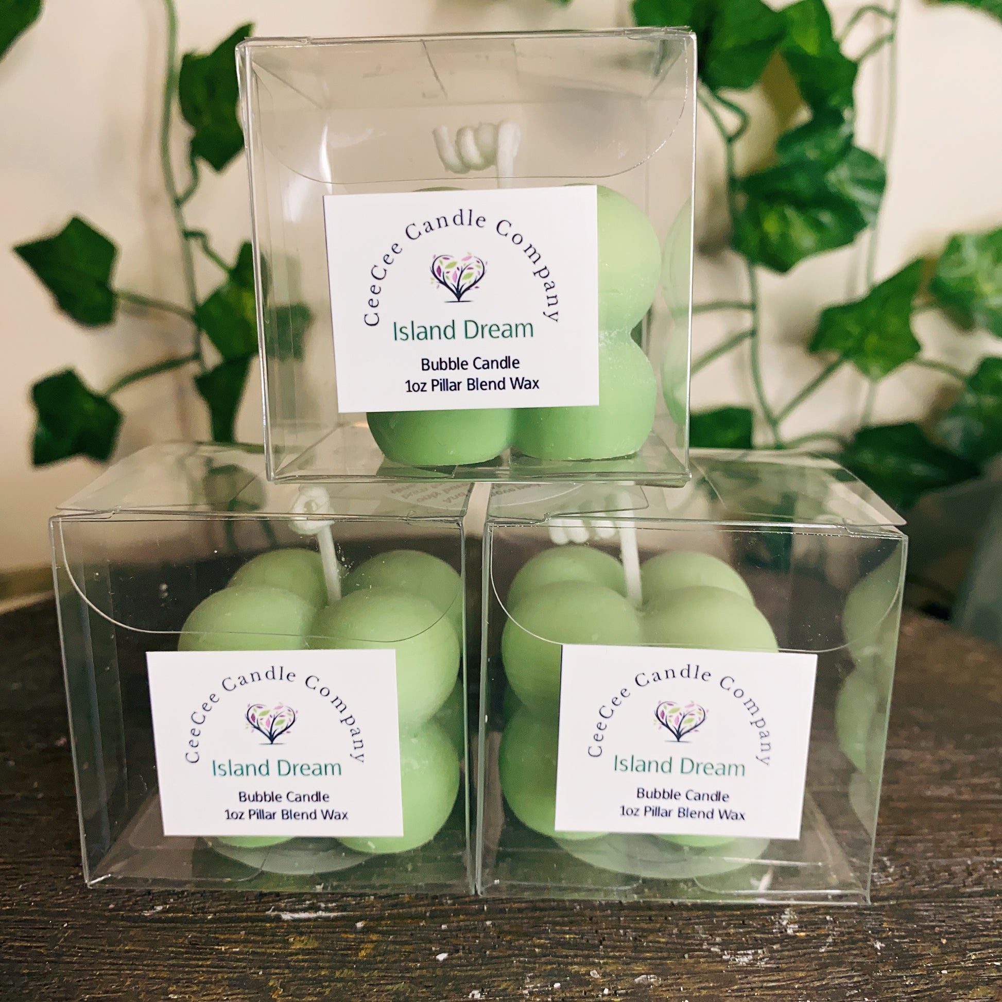 Three light green Bubble Candles with glass dishes from CeeCee Candle Company are stacked in a pyramid. Each candle adds lush fragrance, echoing the blurred green leaves in the background.