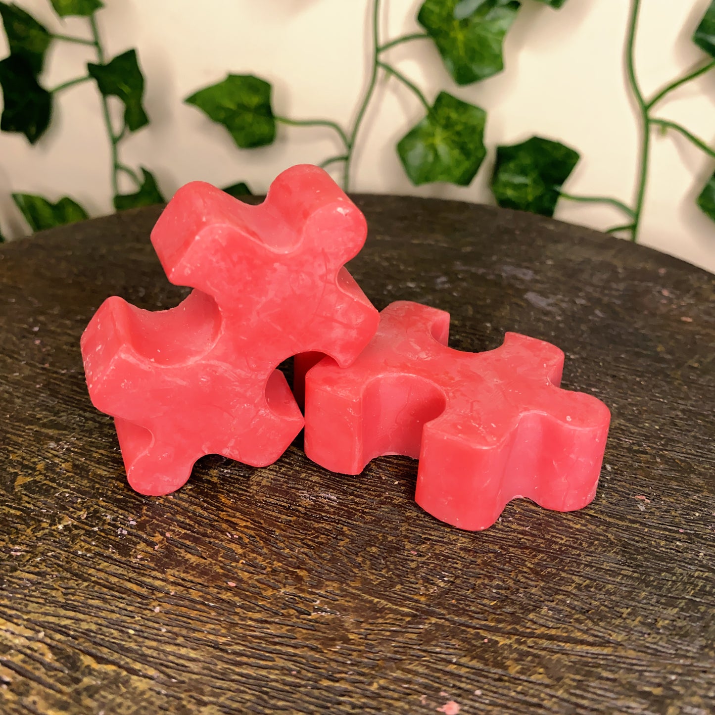 Three red, puzzle-shaped Puzzle Pieces Wax Melts from CeeCee Candle Company are stacked on a dark wooden surface, with green ivy leaves blurred in the background.