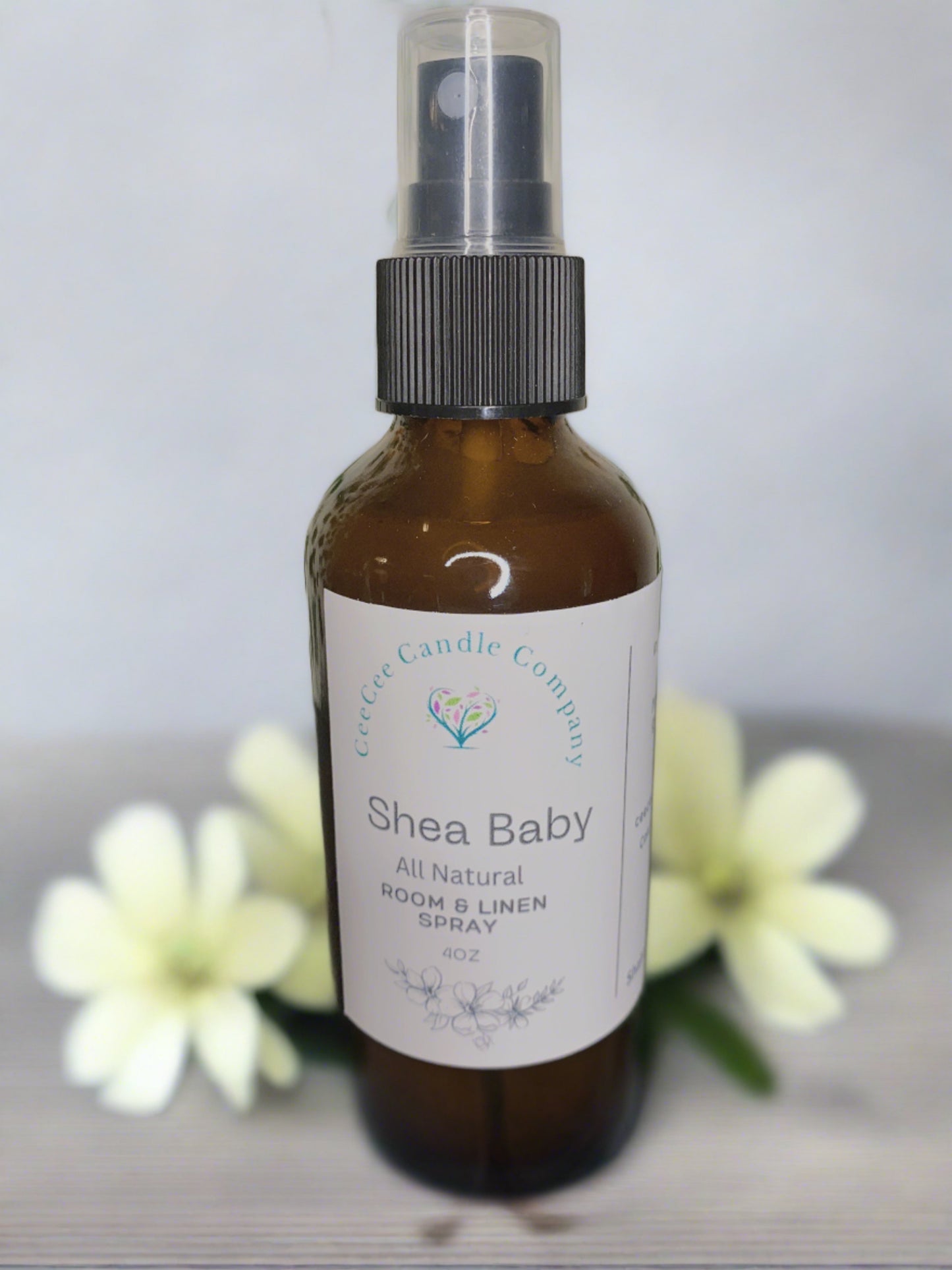 A brown bottle of CeeCee Candle Company Room & Linen Spray – Clean, Long-Lasting Scents is shown, with the label visible and soft white flowers blurred in the background.