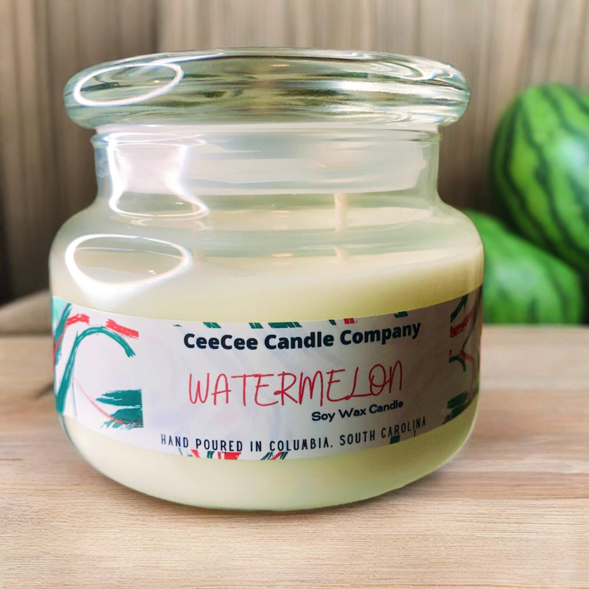A glass jar Double Wicked Apothecary Soy Candle by CeeCee Candle Company sits on a wooden surface, with a blurred watermelon in the background. The label notes it is hand-poured in Columbia, South Carolina.