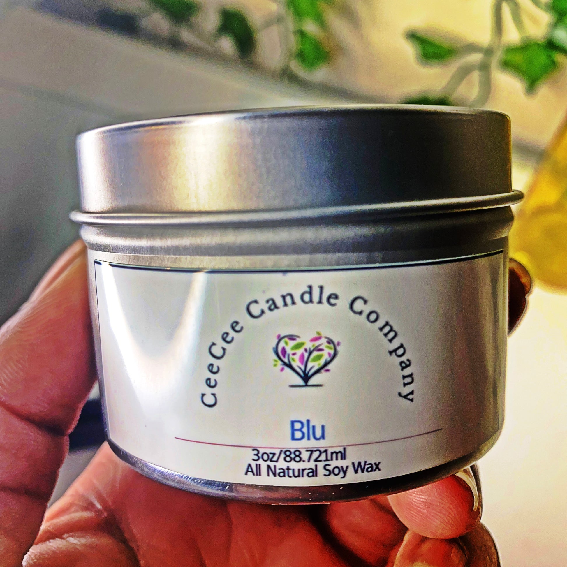 A hand holds a CeeCee Candle Company Travel Size Soy Candle in a silver tin, featuring a white label with tree logo; blurred greenery highlights its mood-boosting fragrance.