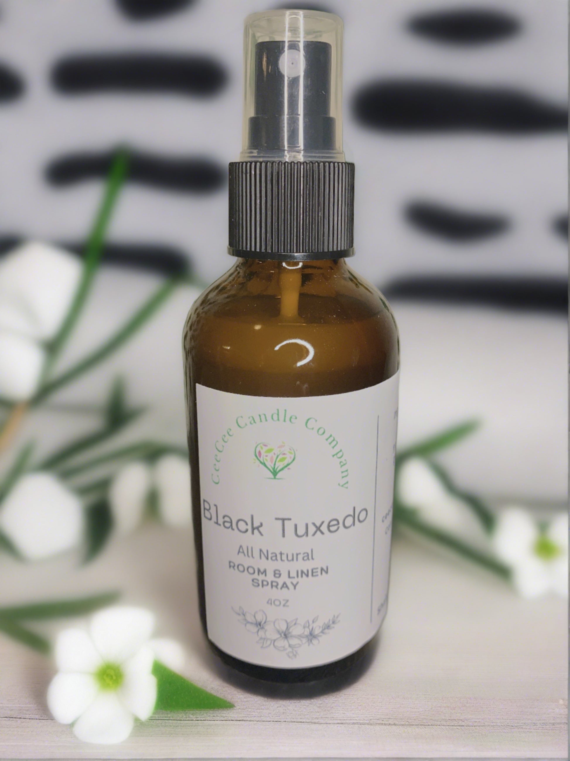 A brown glass bottle labeled “CeeCee Candle Company Room & Linen Spray – Clean, Long-Lasting Scents” is surrounded by green leaves and white flowers, emphasizing signature scents against a blurred black and white background.