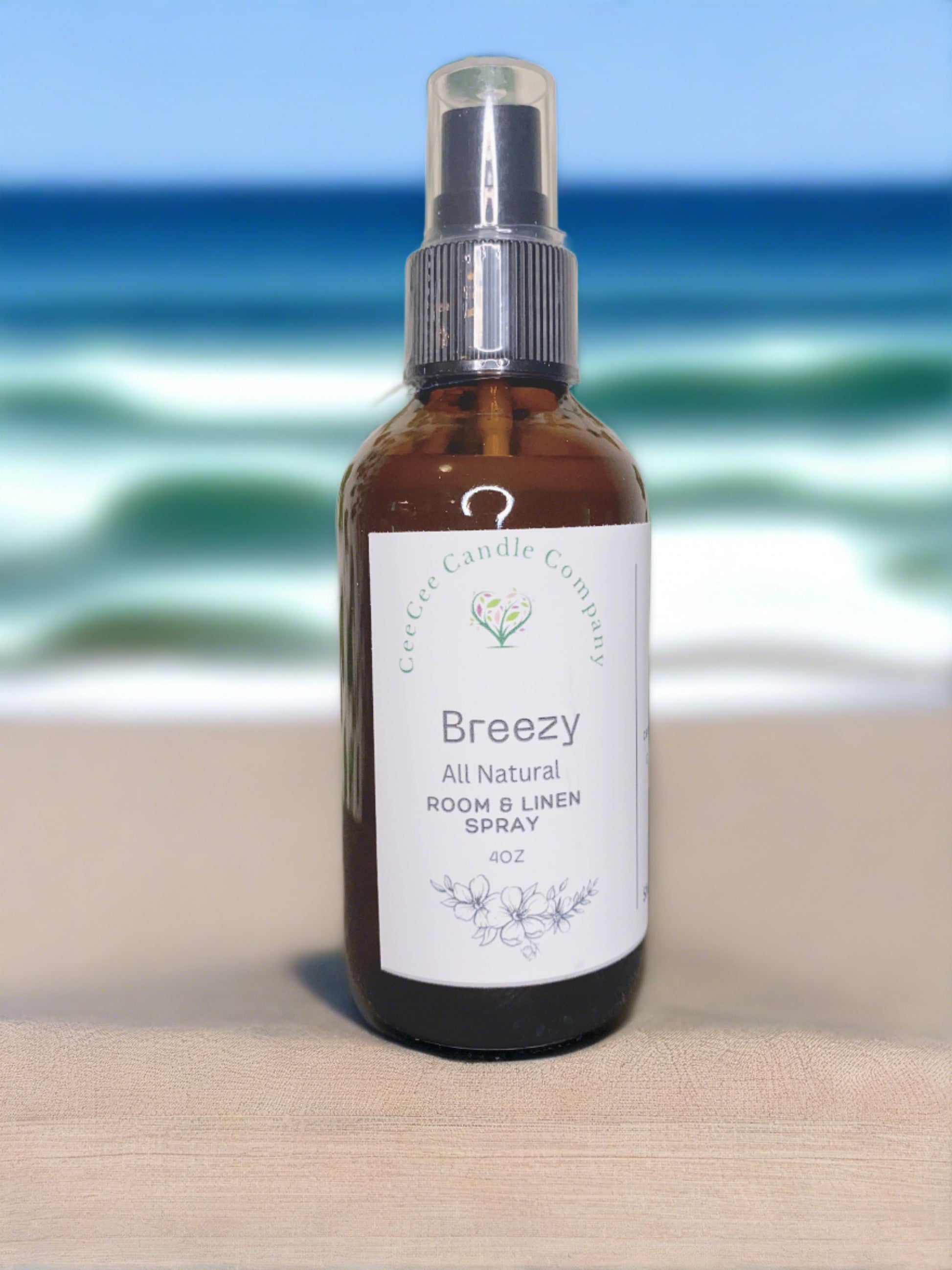 A brown glass spray bottle labeled “CeeCee Candle Company Room & Linen Spray – Clean, Long-Lasting Scents” stands on sandy ground, capturing the essence of CeeCee Candle Company against a blurred ocean and sky backdrop.