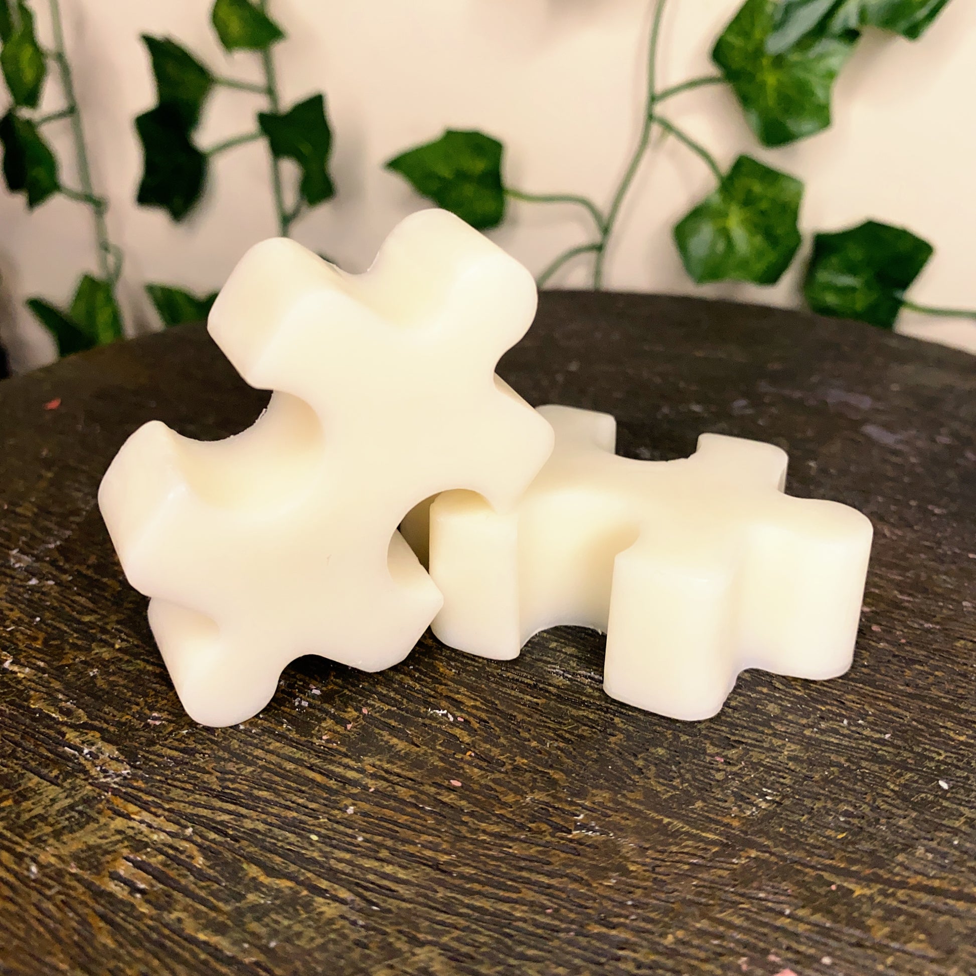 Two white Puzzle Pieces Wax Melts by CeeCee Candle Company sit on a dark wooden surface, with softly blurred green leafy vines in the background.