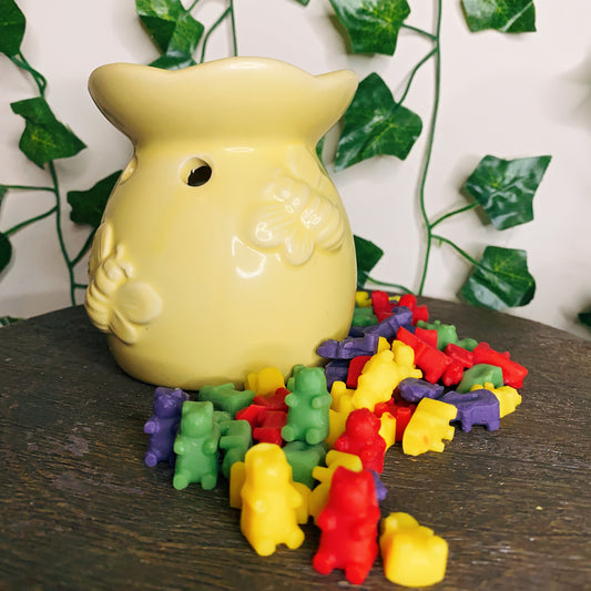 CeeCee Candle Company's Moroccan Cashmere Gummy Bear Wax Melts in red, yellow, green, and purple surround a yellow ceramic pot on a wooden surface, with green ivy leaves adding a cozy touch to the scene.