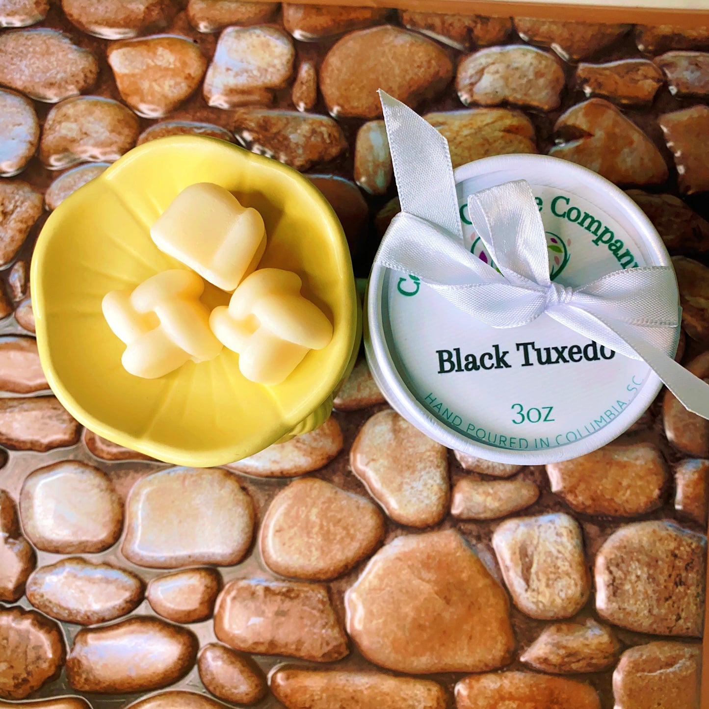 A yellow bowl with four heart-shaped wax melts from the Wax Melts Gift Box by CeeCee Candle Company sits beside a "Black Tuxedo, 3oz" tin on a stone-patterned surface, with the tin tied using a white ribbon.