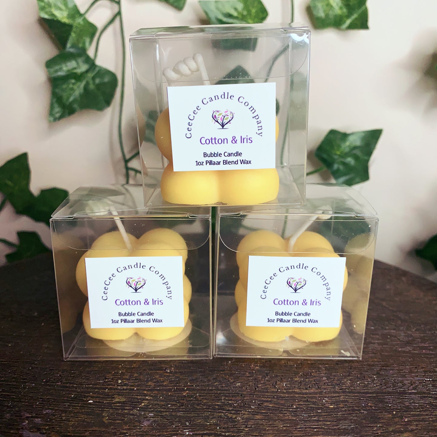 Three yellow Bubble Candles with glass dishes by CeeCee Candle Company are stacked on a dark wooden surface. Each is labeled "Bubble Candle," ideal for pairing with your favorite fragrance oils. Green leaves appear in the background.